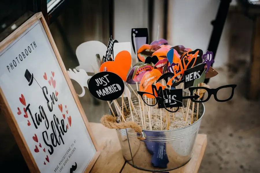Accessoires de photobooth sur bâtonnets réunis dans un seau : lunettes, moustaches et pancartes, à côté d'un panneau selfie