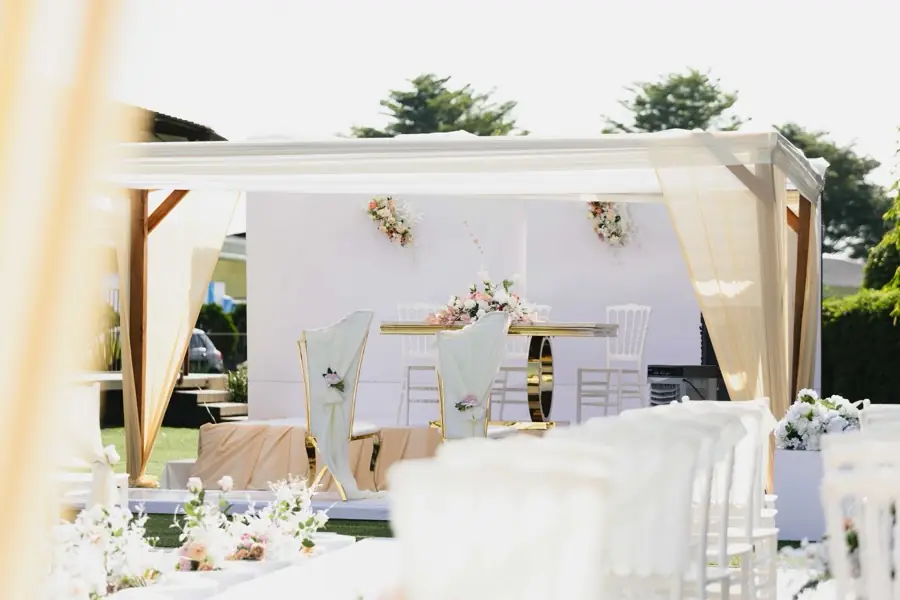 Coin photo de mariage en extérieur : panneau blanc décoré de compositions florales sous une tonnelle aux voilages clairs